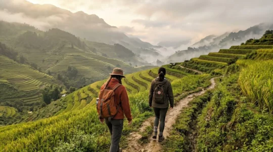 Deux voyageurs vus de dos marchent sur un sentier étroit entre les rizières en terrasses de Sapa sous une brume matinale légère